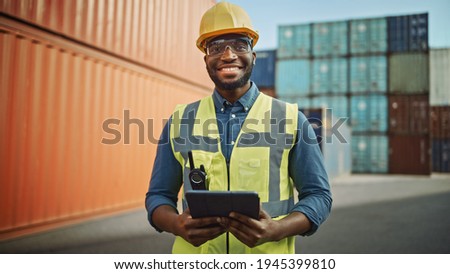 Smiling Portrait of a Handsome African American Black Industrial Engineer in Yellow Hard Hat and Safety Vest Working on Tablet Computer. Foreman or Supervisor in Container Terminal. Royalty-Free Stock Photo #1945399810