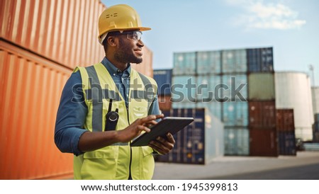 Smiling Portrait of a Handsome African American Black Industrial Engineer in Yellow Hard Hat and Safety Vest Working on Tablet Computer. Foreman or Supervisor in Container Terminal. Royalty-Free Stock Photo #1945399813