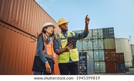 Multiethnic Female Industrial Engineer with Tablet and Black African American Male Supervisor in Hard Hats and Safety Vests Stand in Container Terminal. Colleagues Talk About Logistics Operations. Royalty-Free Stock Photo #1945399819