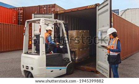 Forklift Driver Loading a Shipping Cargo Container with a Full Pallet with Boxes in Logistics Port Terminal. Latin Female Industrial Supervisor and Safety Inspector with Tablet Managing the Process. Royalty-Free Stock Photo #1945399882