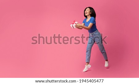 Happy Holiday. Full body length shot of cheerful asian woman holding gift box looking away and giving present to the side to free copy space, jumping isolated on bright color pink studio background