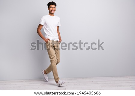 Full size photo of brunet optimistic curly guy go wear white t-shirt trousers sneakers isolated on grey color background