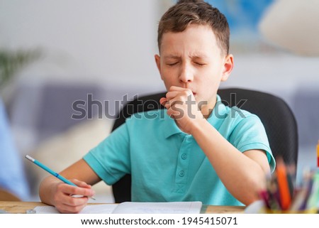 small child, schoolboy of 8 years old, is studying at a desk with laptop, at home. School distance education at home during quarantine concept. Coughing, sneezing, covering your mouth with your hand. Royalty-Free Stock Photo #1945415071