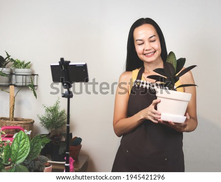 Asian female gardener in  sleeveless shirt and apron, working at home,  using mobile phone to live or record video selling her houseplant or presenting how to take care plant.  selective focus.