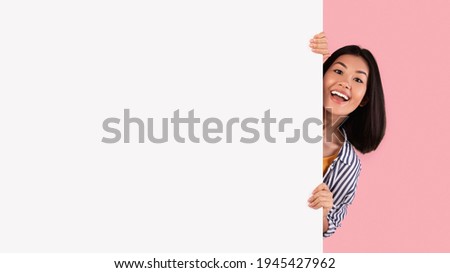 Intresting Offer. Happy Casual Asian Woman Peeping Out The Side Of White Advertisement Board For Your Text Or Design. Smiling Lady Holding Billboard, Looking At Camera, Standing Over Pink Wall