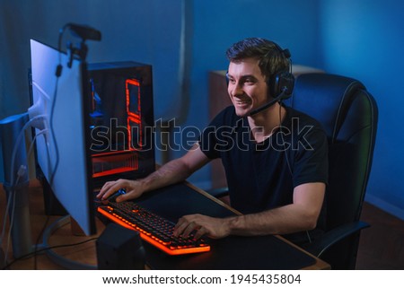 Young professional gamer playing online video games on his PC. He is smiling because of winning the game, having live stream for his followers and subscribers. Cyber sport concept. Team play Royalty-Free Stock Photo #1945435804