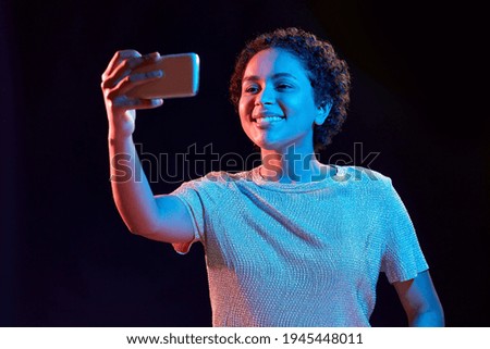 nightlife, technology and people concept - happy young african american woman taking selfie with smartphone in neon lights over black background