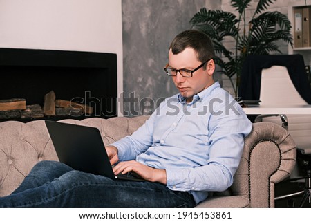 Young successful man in a shirt and glasses works with a laptop while sitting in a large spacious office on the couch against the background of the fireplace. Success and time management concept.