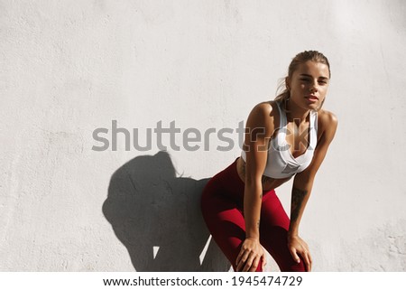 Sweaty fitness girl taking breath on morning jogging. Female athlete runner resting, workout outdoors on fresh air, jogger take break, wearing sports outfit, standing near concrete wall Royalty-Free Stock Photo #1945474729