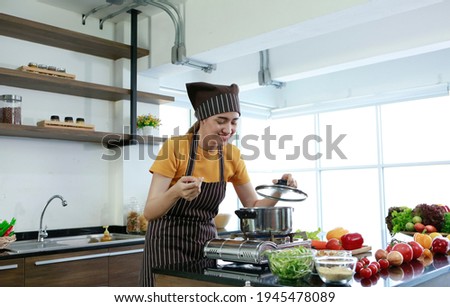 Beautiful Asian women smiling standing smell soup from pot. Housewife chef with apron cooking meat steak and salad in house kitchen for sell food box online and delivery. Work from home covid concept
