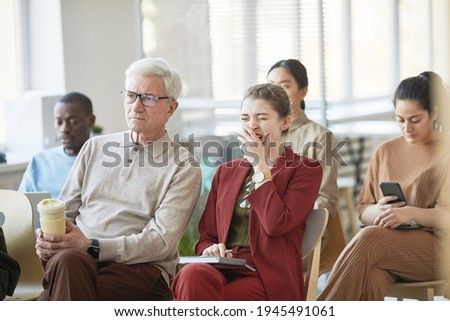 Diverse group of business people sitting on chairs in audience and listening at meeting or seminar, focus on young businesswoman yawning in foreground