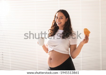 Beautiful pregnant woman holding croissant and cup of coffee in her hands during morning breakfast. Concept of good health and positive attitude while expecting baby