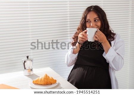 Joyful pregnant young beautiful woman looks through blinds during her morning breakfast with coffee and croissants. Concept of good morning and waiting for meeting with a baby. Copyspace