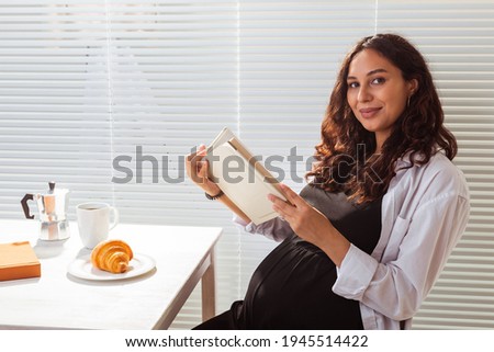 Side view of happy young beautiful woman reading book while having morning breakfast with coffee and croissants on background of blinds. Good morning concept and pleasant lunch break