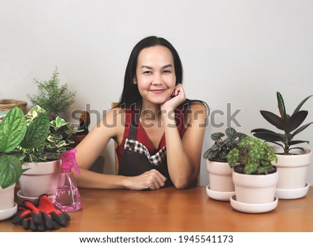 Happy Asian female gardener in  sleeveless shirt and apron, working at home, sitting with her houseplants and gardening tools  indoor, chin on hand, smiling and looking at camera. gardening concept.