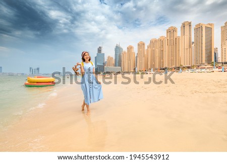 Cheerful woman takes off her sandals and walks barefoot on the water on the beach in Dubai at the Jumeirah Beach residence. The concept of travel and vacation in the UAE Royalty-Free Stock Photo #1945543912