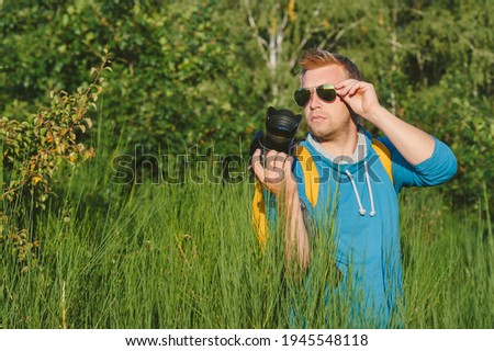 Male Tourist, straightens his glasses and holds a professional photo-video camera in his hands. Against the backdrop of beautiful nature
