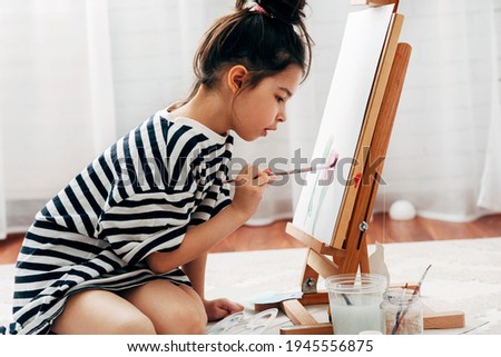 Side view of a little girl painting with a paintbrush on the paper on an easel at home. A cute kid sitting on the floor and drawing in her room during the lockdown.