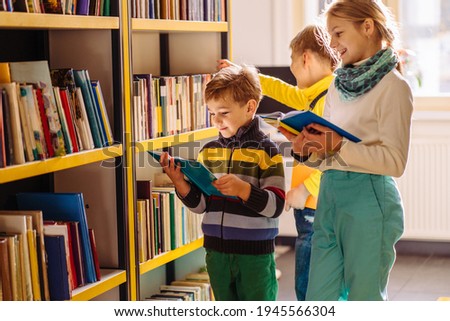 Cute different age pupils, two boys and girl looking for books in library at the elementary school. Three children standing near bookshelves and reading together at school's library.