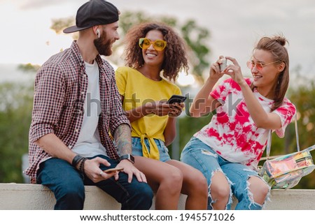 colorful stylish happy young company of friends sitting park, man and women having fun together, summer hipster fashion style, traveling with camera, taking selfie pictures, smiling and positive