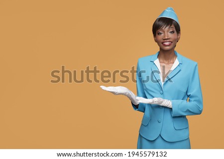 Smiling young african stewardess shows a welcome gesture. Orange background. Royalty-Free Stock Photo #1945579132