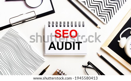 On a light table are notebooks, a magnifying glass, an alarm clock, glasses, and a pen. And in the center is a notebook with the text SEO AUDIT