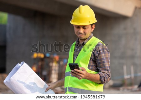 Asian construction workers using smartphone and holding blueprint at the construction site Royalty-Free Stock Photo #1945581679
