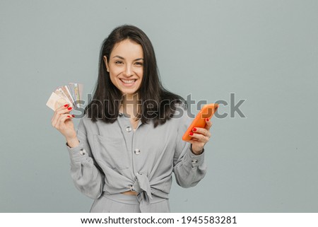 Photo of cute smiling female holds smartphone and credit cads. Wears grey t-shirt, isolated grey color background.