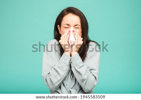 Photo of sick female is sneezing. Wears grey shirt, isolated turquoise color background.