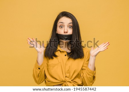 Concept photo of female victim with her mouth taped up, holds her head. Wears yellow shirt, isolated yellow color background.