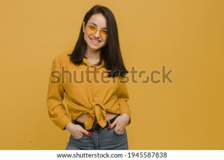 Photo of young female wears sunglasses and looks so kind and happy. Wears yellow shirt, isolated yellow color background.