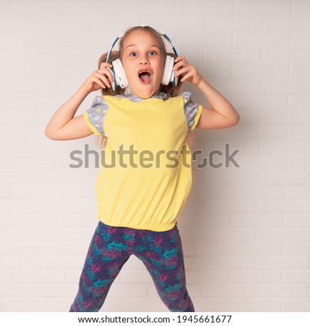 little excited girl listens to music on headphones. She dances and jumps merrily to the music in blue leggings and a yellow T-shirt against the backdrop of a white brick wall