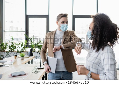 interracial business partners in medical masks doing elbow greeting in office Royalty-Free Stock Photo #1945678510