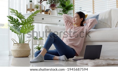 Young attractive beautiful asia female girl or university student sit smile look outside window put arm hand back behind head at sofa couch living room feeling relax comfort at cozy home houseplant. Royalty-Free Stock Photo #1945687594