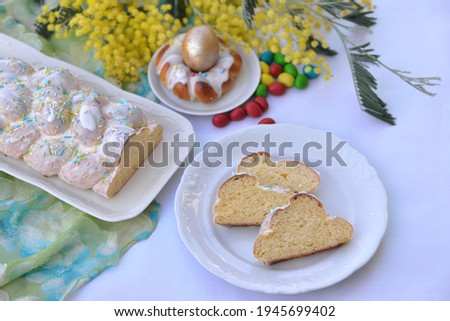Table with Easter food: sweet braided bread with sugar glaze and bread texture of bread slices on another plate, Easter cake in a shape of Easter nest with an egg; mimosa flowers in background