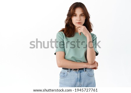 Young thoughtful lady looking at camera pensive, touching chin and thinking, standing in t-shirt and jeans against white background