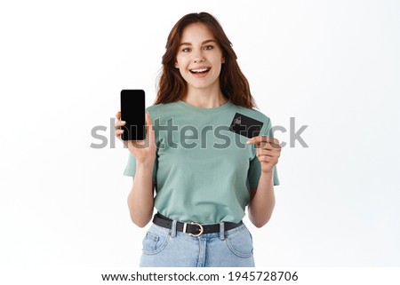 Portrait of a happy young brunette girl showing plastic credit card while holding mobile phone isolated over white background