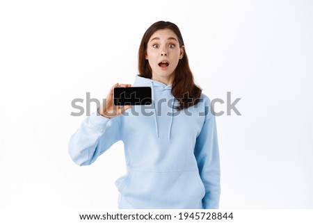 Excited teenage girl showing application or video game on empty mobile phone screen, demonstrates your promo on smartphone display, standing against white background
