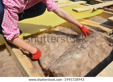 Roofing construction: A DIYer, a woman is insulating the roof with mineral wool, mineral glass batt placing insulation material between the ceiling joists of the roof.  Royalty-Free Stock Photo #1945728943