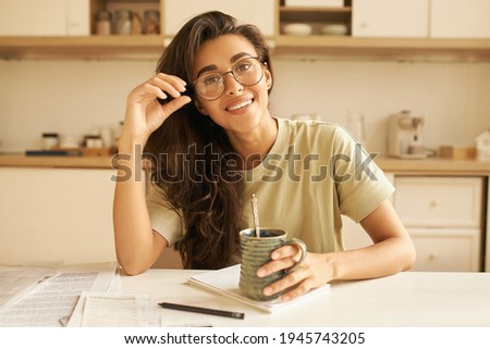 Cute student girl wearing stylish glasses having tea in kitchen, sitting at dining table with mug, copybook and sheets of paper, doing homework. Attractive young Arabic female relaxing at home