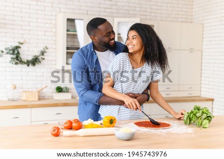 Overjoyed African American couple in love standing next to kitchen counter, hugging, spending leisure time weekend together at home, looking at each other, cooking, girl is spreading sauce on pizza