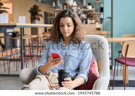 Hispanic latin young woman student holding smartphone and coffee sitting in modern space studying on cell, browsing, checking apps, texting using mobile phone in university campus or creative office.