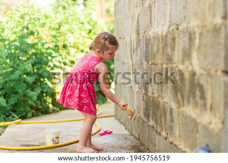 
A little girl in a dress with watermelons paints the wall with a brush. Summer outdoor activities. Paints a brick wall with water.