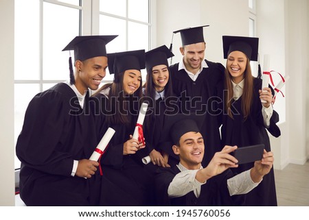 Selfie for memories. Group of happy international students in mortar boards and bachelor gowns with diplomas taking selfie using smartphone in classroom. Graduation, technology and people concept.