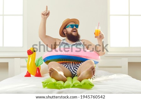 Funny guy in sunglasses, sun hat and swim ring sitting on beach mattress and sipping cocktail points finger up struck by cool idea on how to turn quarantine holiday at home into fun summer vacation Royalty-Free Stock Photo #1945760542