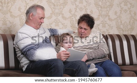 Grandfather, grandmother are playing with toddler together on modern gadget in room. Child, girl shows something in tablet to grandparents. An elderly couple with granddaughter are using tablet online