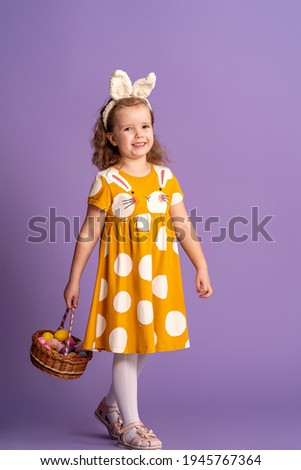charming smiling little girl in full height walks on purple background in studio and holds basket Easter eggs in her hands. Funny happy baby wears rabbit ears on Easter day. Copy space.