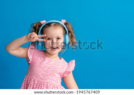 positive little girl with headphones looks into frame, listening to music on blue background. Copy space.Full emotion. Enjoying song playing in headphones. child sings and dances, big music lover.