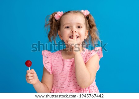 Close-up of a funny child showing a finger gesture to his lips. naughty little girl with a lollipop on a blue background in the studio posing with pleasure. Copy the space.