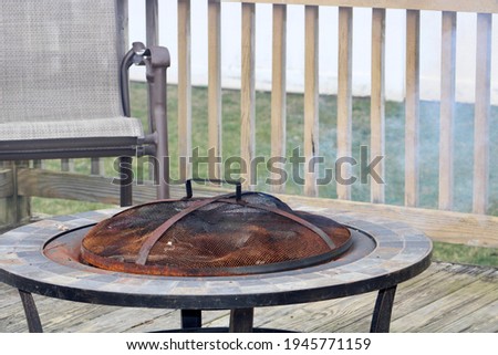 A rod iron fire pit with green tiles in a circle shape. The fire place is sitting on a wooden deck in front of two empty lawn chairs. There are charred logs under the top that are smoking and on fire.